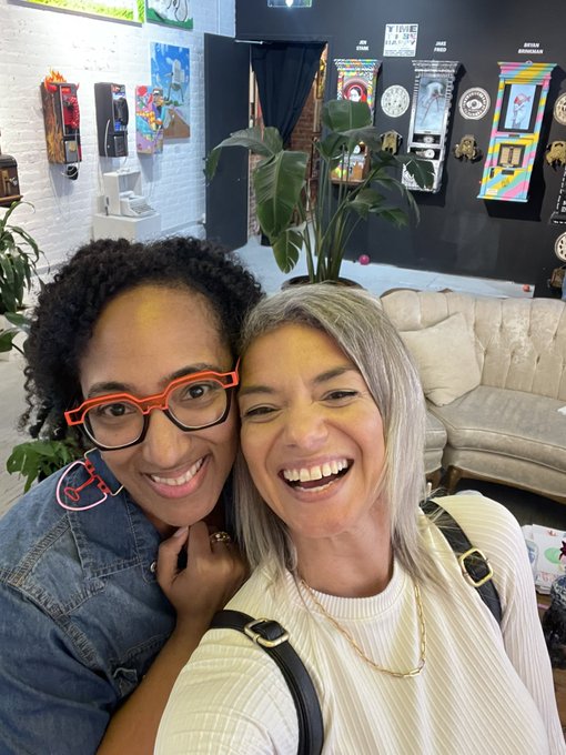 Two smiling women pose close together for a selfie inside what appears to be a quirky shop, with various clocks and eccentric items mounted on the wall behind them.