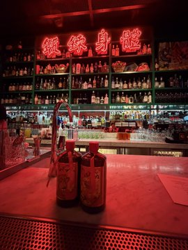 A dimly lit bar bathed in red neon glow, featuring two bottles in the foreground and a backlit shelf of liquor bottles beneath a Chinese neon sign, evoking the underground, intimate atmosphere of a BAYC meetup in Hong Kong.