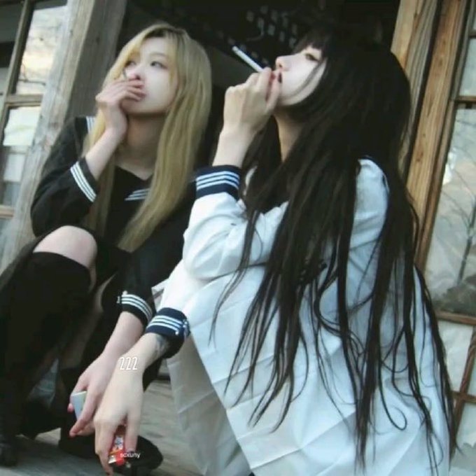 Two young women pose in matching varsity-style jackets against a wooden background, one in black and the other in white, both with long flowing hair—one blonde, one dark—while exhaling smoke, capturing a rebellious aesthetic that mirrors the post's themes of retaliation and rivalry.