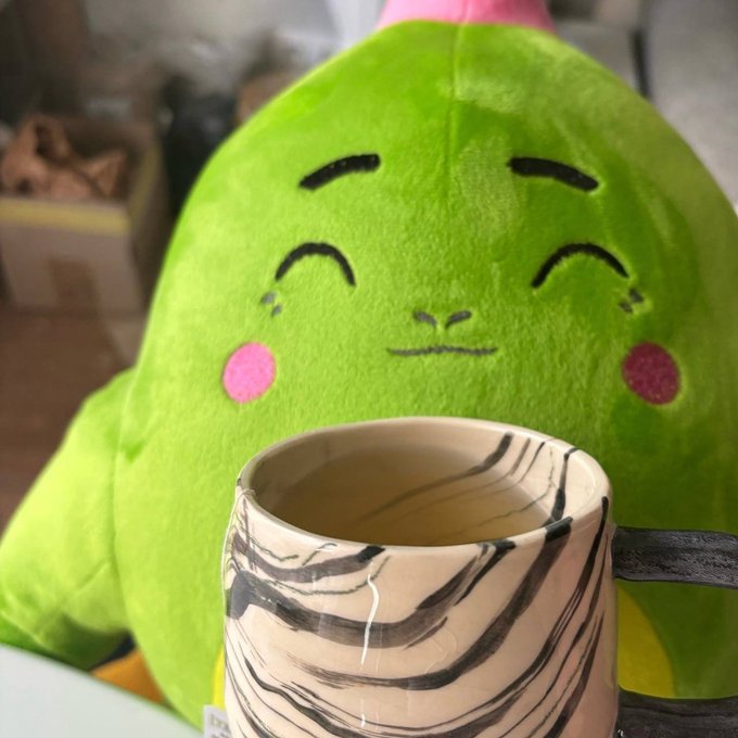 The image features a green stuffed animal with a cup of coffee placed next to it. The stuffed animal appears to be a cute plush toy, possibly a teddy bear or a frog, sitting on top of a table. The cup of coffee is positioned in front of the stuffed animal, creating an interesting contrast between the softness of the toy and the warmth of the beverage.