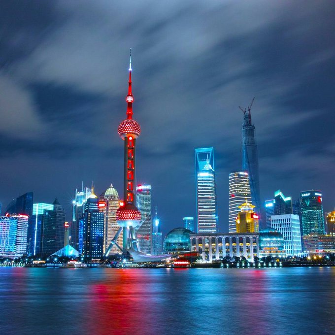 A nighttime cityscape of Shanghai's Pudong skyline, prominently featuring the Oriental Pearl Tower illuminated in red against a dark, cloudy sky, with reflections on the water.