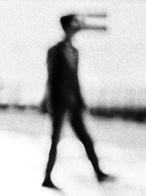 A blurry, grainy black-and-white artistic image showing a shadowy figure of a person walking, creating an eerie silhouette effect.