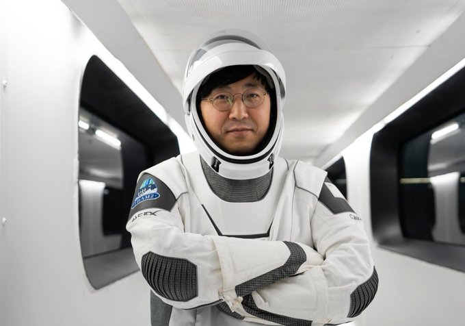 The image features a man dressed as an astronaut, standing in front of a white wall with his arms crossed. He appears to be posing for a photo or possibly giving a speech at an event related to space exploration. The background includes several windows that provide natural light and a view of the sky outside. There is also a reference to Bitcoin in the image, which could indicate that this astronaut might have some knowledge about cryptocurrency or its potential applications in space travel.