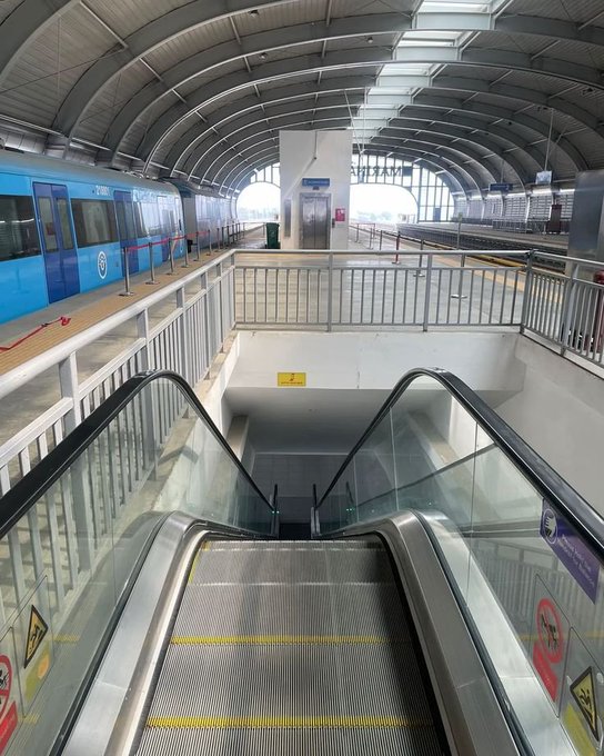 A view looking down an escalator at a modern train station, with a blue and white train parked on the left platform under a curved, industrial-style roof.