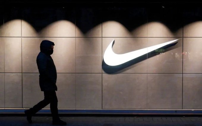 A nighttime photograph of a pedestrian in a hooded jacket walking past an illuminated Nike swoosh logo displayed on a tiled wall.