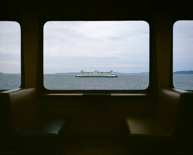 A view through rounded windows from inside a ferry, looking across the sea at a large white passenger ferry boat under an overcast sky.