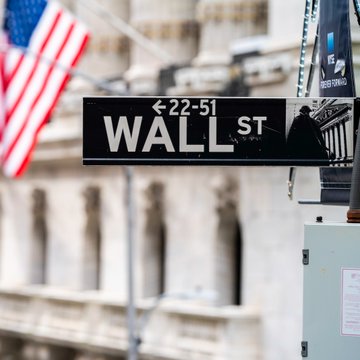 The image shows a street scene featuring a black and white sign for "Wall St", indicating that this location is associated with the financial district of New York City. The sign also displays an arrow pointing towards the Wall Street Financial Center, which is a prominent landmark in the area. In the background, there are two American flags flying high on poles, symbolizing patriotism and representing the United States' role as a global economic power. The image also includes text that references Bitcoin ETFs and the identity crisis they have caused within the financial system.