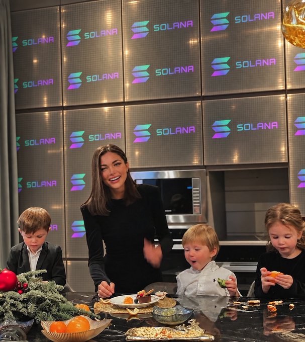 A woman in a black dress smiles alongside three children at a table with food, with multiple Solana logos displayed on screens in the background.