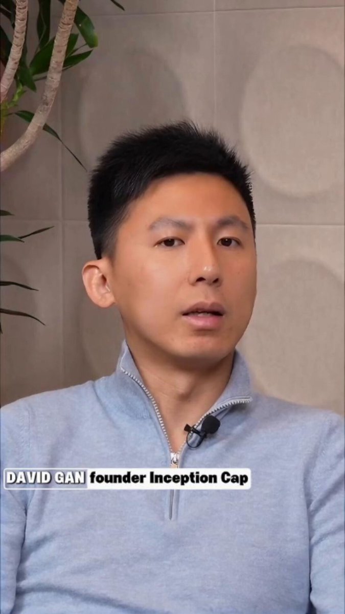 The image features a man wearing a blue sweater, sitting against a wall with his head tilted slightly to one side as he looks at someone or something off camera. The background includes a plant behind him, adding some greenery to the scene. The main focus of the image is on the man and his facial expression, which suggests that he might be engaged in a conversation or discussing an interesting topic.