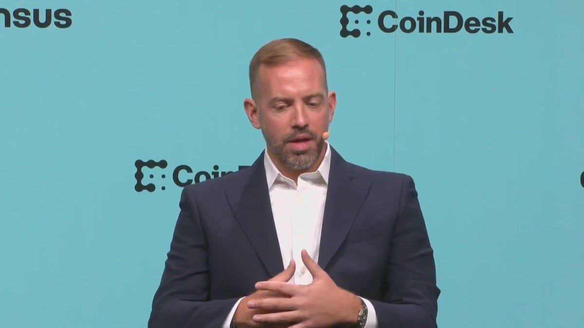 The image features a man dressed in a suit, standing in front of a blue wall with the words "CoinDesk" written on it. The man appears to be giving a speech or presentation, as he has his hands together in front of him. The background includes logos and text related to cryptocurrency and digital currency, suggesting that this event is connected to the world of blockchain technology and financial innovation.