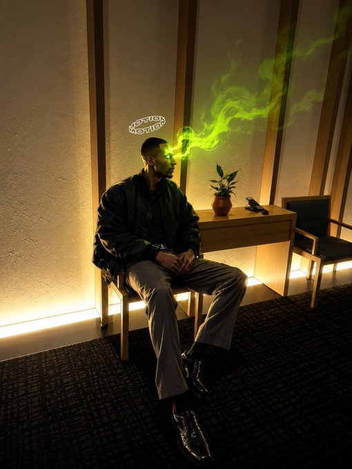 A young man sits on a chair in a dimly lit room, exhaling green smoke while wearing a small glowing halo that reads 'MEXICAN MAFIA' above his head.