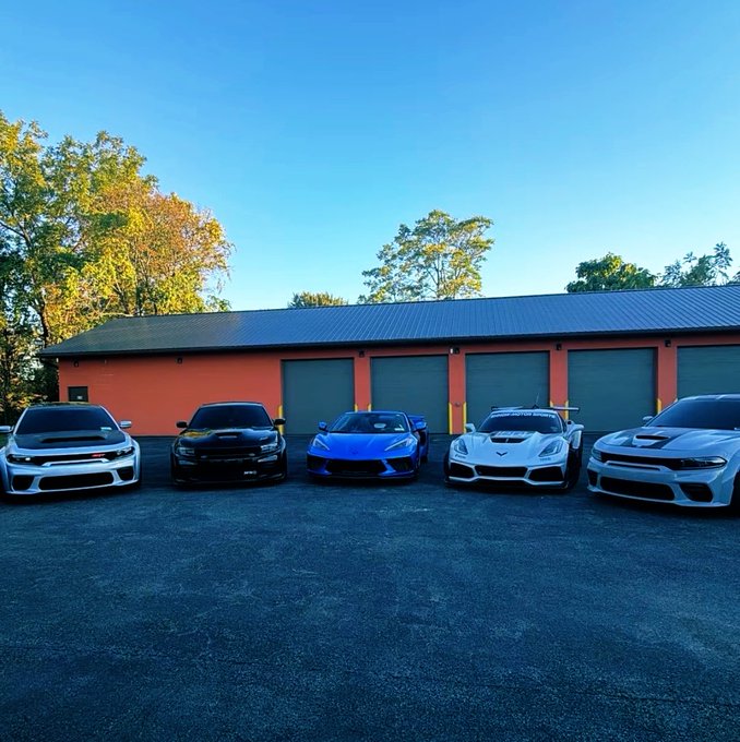 A lineup of five performance cars (including a Chevrolet Camaro, Dodge Charger, Toyota Supra, Corvette, and Dodge Charger) parked in front of a red building with multiple garage doors.