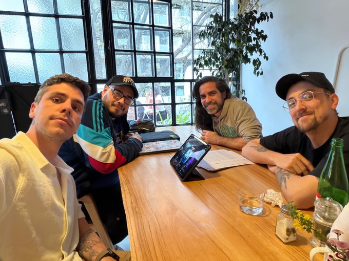 Four men sit around a wooden table in a sunlit room with large windows, collaborating over a tablet displaying a game design, with the @lcbgamestudio branding on one man's jacket and a small potted plant and water glass on the table. The scene captures a creative work session tied to the post about building new games and designing a fresh pixel logo for @ethlambda_lean.