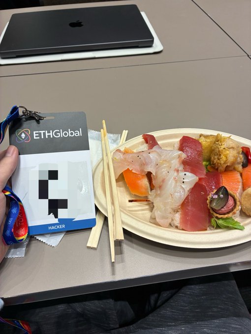 A hand holds an "ETHGlobal HACKER" badge next to a plate of assorted sushi and chopsticks, with a closed laptop visible on the gray table in the background.