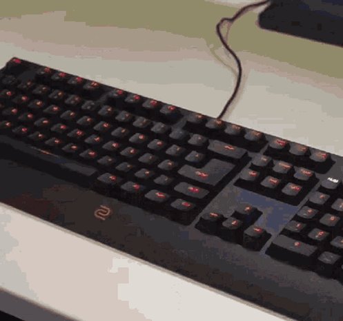 The image shows a black computer keyboard with red keys, placed on top of a white desk. The keyboard appears to be new or recently used, as there are no visible signs of wear or damage. The background of the image is blurred, drawing attention to the keyboard and its surroundings.