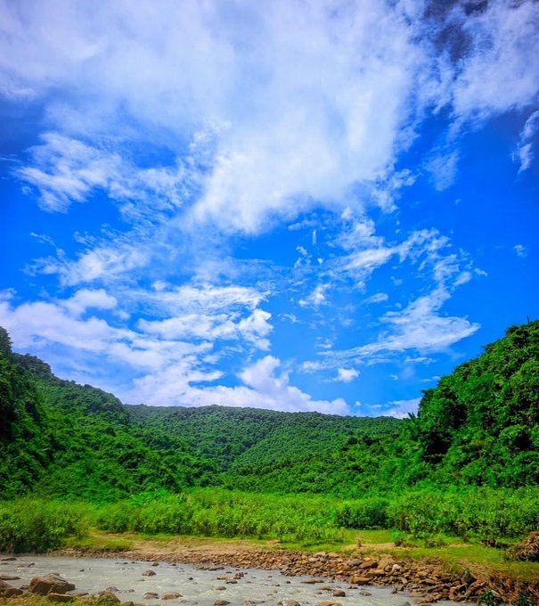 A scenic landscape photograph featuring a vibrant blue sky with scattered clouds above lush green forested hills and a rocky stream in the foreground.