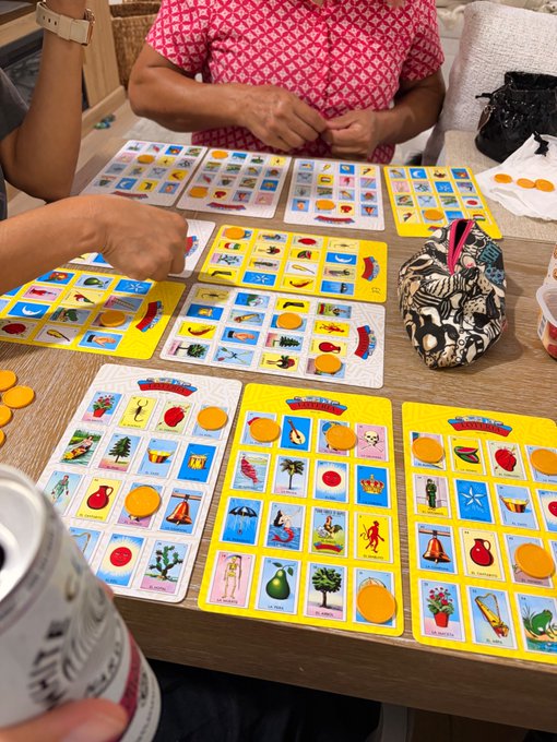 Two people sit at a table playing a game of Loto (bingo), covering pictures on yellow cards with orange chips while holding drinks.