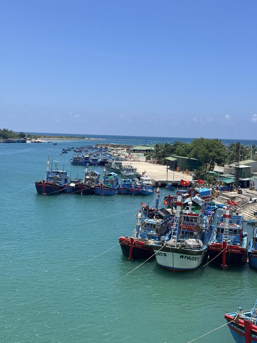 An aerial view of a harbor filled with numerous fishing boats docked along a pier, with turquoise waters, sandy beaches, and a clear blue sky in the background.