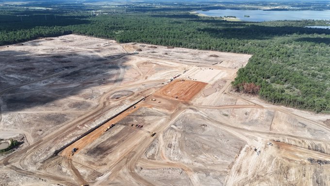An aerial view reveals a sprawling construction site where heavy machinery has cleared massive swathes of forest, transforming the landscape into a graded expanse of dirt and sand marked with tire tracks and construction zones. The image visually supports the post's claim about "Terafab" by showing the rapid, aggressive land-clearing progress at the site.