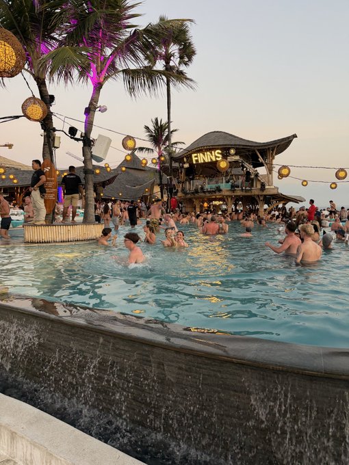 A lively beach club pool party at dusk, featuring a large swimming pool with floating patrons, palm trees with hanging lights, and a bar structure displaying the name 'FINNS'.