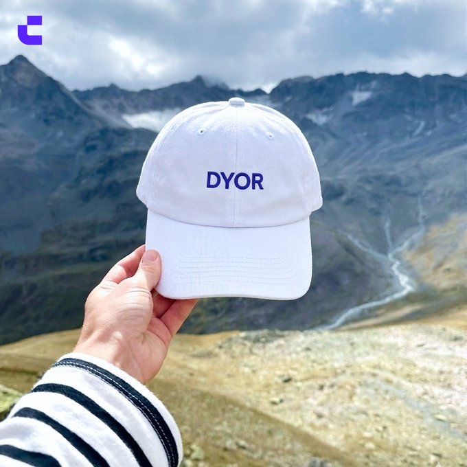 A hand holds up a white baseball cap embroidered with "DYOR" (Do Your Own Research) against a mountainous backdrop, cleverly reinforcing the post's message that everyone should research before investing in any cryptocurrency.
