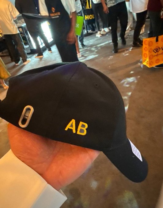 A black cap with "AB" embroidered in yellow is held in someone's hand at what appears to be a BNB community event, commemorating a custom cap received from the BNB team in Dubai. The yellow lettering directly reflects the post's message about supporting the community and tokens, with the "AB" referencing the $AB mentioned in the caption.