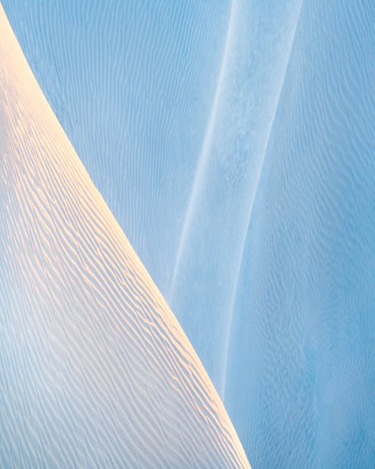 A serene photograph of rippled sand dunes, showing a sharp diagonal ridge line separating warm, peach-toned sand from cooler, blue-shadowed sand.