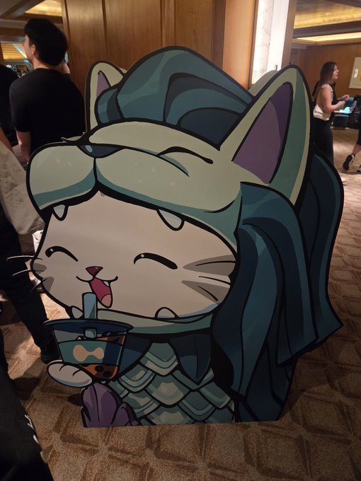 A large cardboard cutout featuring a chibi-style cat character in green armor drinking from a bubble tea cup stands in a conference hallway.
