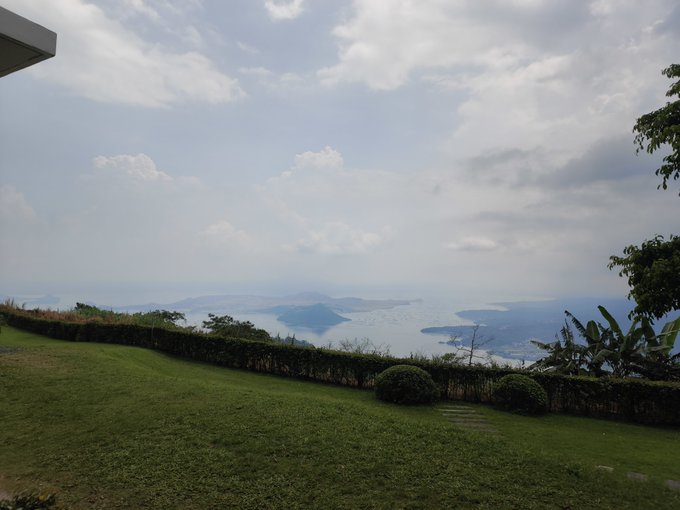 A scenic landscape view featuring a lush green lawn and hedge in the foreground, overlooking a hazy coastal area with islands in the distance under a partly cloudy sky.