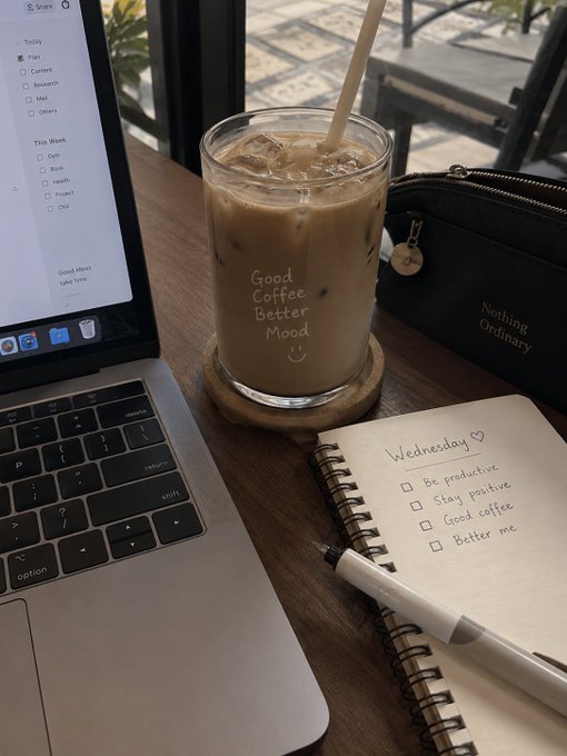 A flat lay of a cafe scene featuring an iced coffee glass labeled 'Good Coffee Better Mood' next to a MacBook, a 'Nothing Ordinary' cosmetic bag, and a notebook with a 'Wednesday' to-do list.