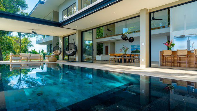 A luxurious modern home features floor-to-ceiling glass walls opening to an indoor-outdoor living space with a swimming pool, hanging egg chairs, and a dining area.