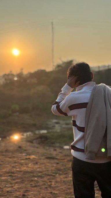 A person in a white and maroon striped sweater watches a sunset over a rural landscape with a utility pole silhouetted against the sky.