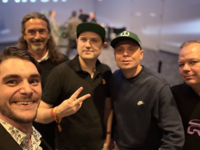 A group selfie of five men at what appears to be a tech event, including one taking the photo with a peace sign, others wearing caps and casual attire like a Nike sweatshirt.