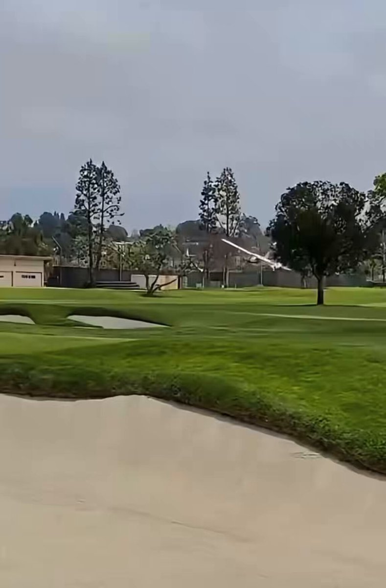 The image shows a golf course with a white golf cart, possibly used by a golfer who has just finished their game. A plane can be seen flying low over the green field, adding an interesting element to the scene. The background features trees and houses, indicating that this is likely a residential area or a golf club.