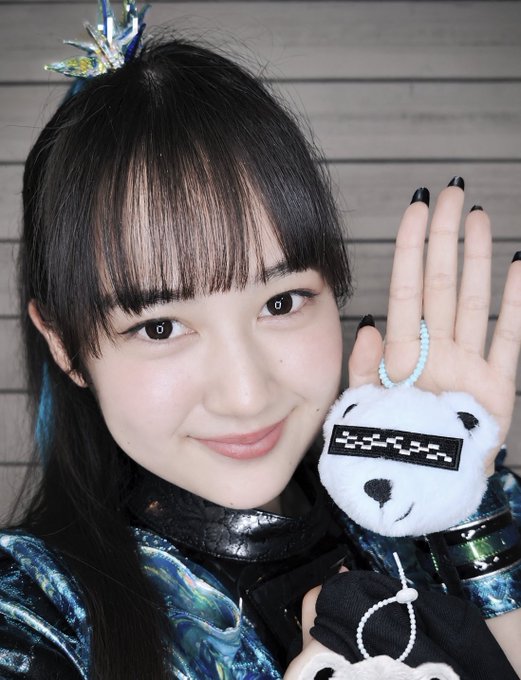 A smiling young woman with long dark hair wearing a shiny teal and black outfit, holding up a small plush bear charm that appears to say "DADA" on it.