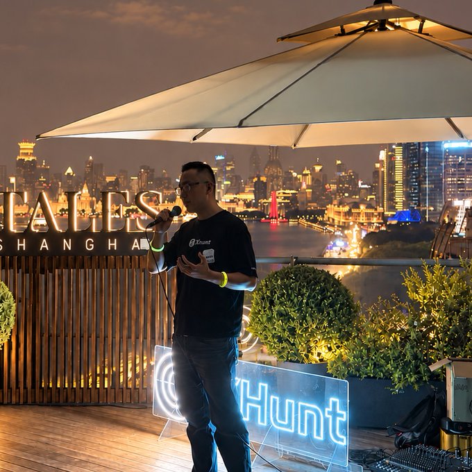 A man holding a microphone speaks at an event with a neon "Hunt" sign, with a Shanghai cityscape and partially visible "SHANGHAI" signage in the background.