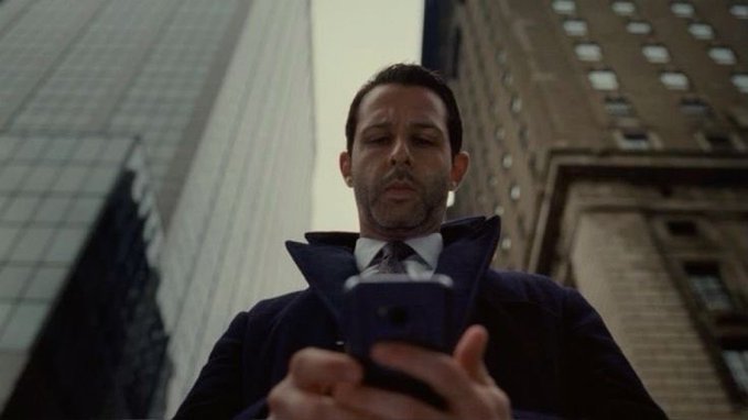 A man in a suit and overcoat stands in an urban setting looking down at his smartphone.