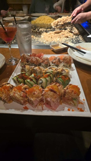 A large platter of assorted sushi rolls topped with sauces and orange roe sits on the table, with a glass of water labeled "EPP" and a pink cocktail nearby, while a hibachi chef prepares food on a grill in the background — a perfect "GM" greeting for a delicious meal ahead.