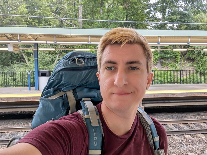 A young man takes a selfie at a train station, wearing a large Osprey backpack, ready for a journey. He is traveling by train to NYC before flying to Berlin to attend the GETDWeb event, with the caption highlighting his upcoming trip sponsored by Zcash and Zcash Community Grants.