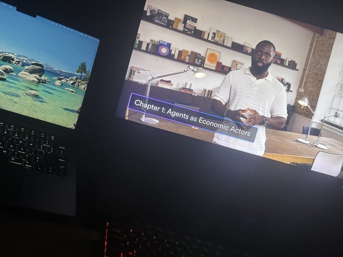 A dimly lit workspace showing a laptop displaying a video lecture titled 'Chapter 1: Agents as Economic Actors' featuring a Black male instructor.