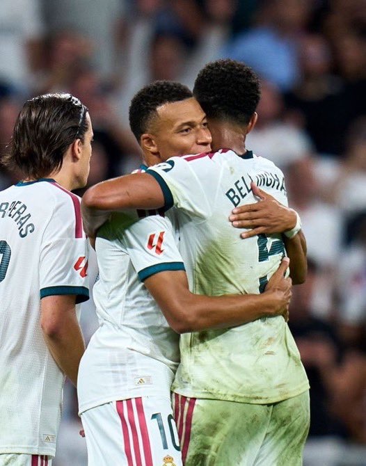 Soccer players in white Real Madrid jerseys embrace in celebration, with the name 'BELLINGHAM' visible on one player's back and 'Mbappé' partially visible on another.