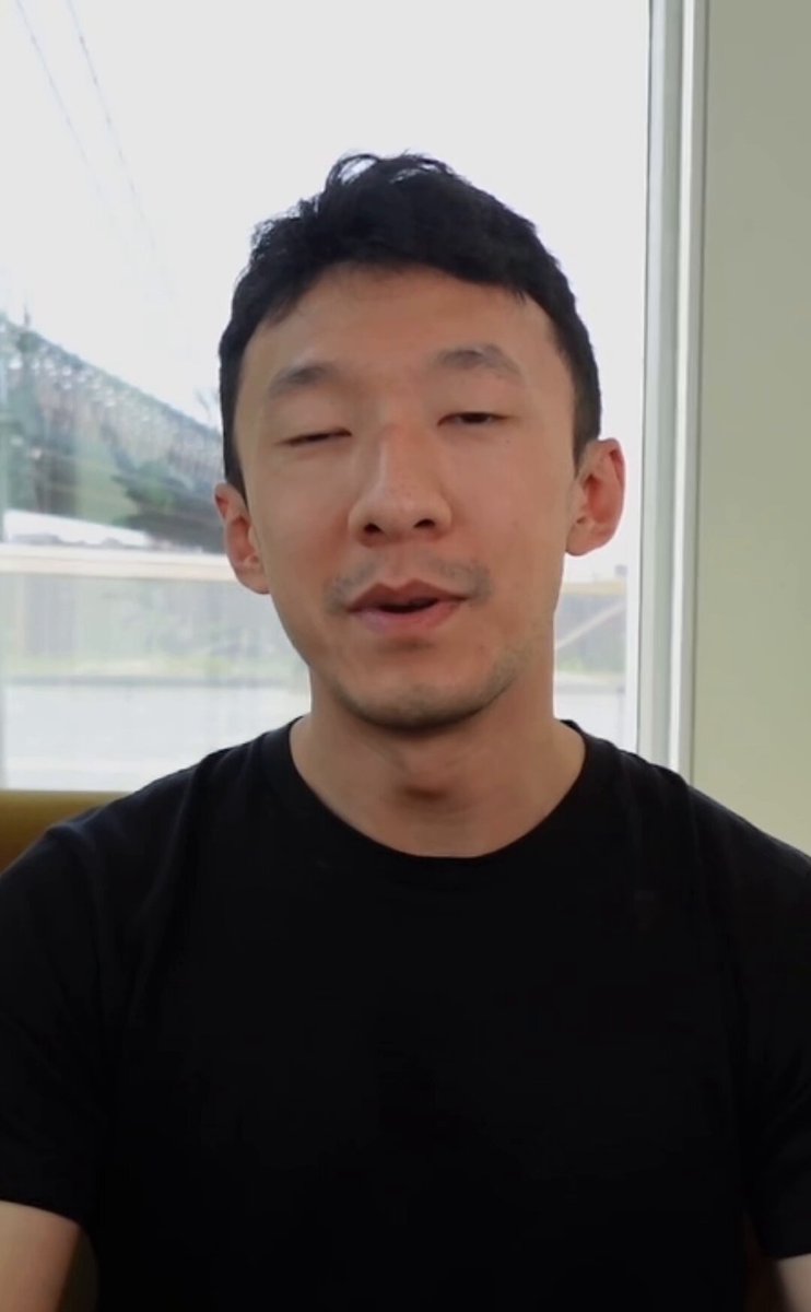 The image features a man wearing a black t-shirt with his eyes closed. The background of the photo shows a window that provides natural light into the room. There are no other objects or people visible in the image, making the focus on the man's face and expression the primary subject matter.
