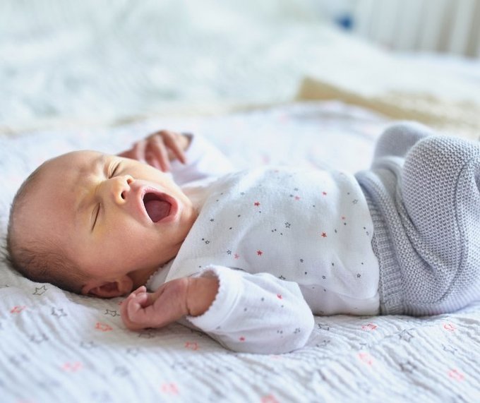 A newborn baby yawns widely while lying on a patterned blanket, wearing a white onesie with small star prints and gray knitted pants.