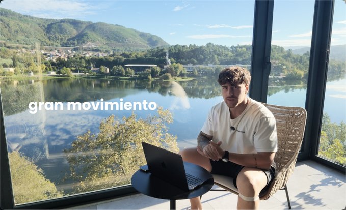 A man sitting in a chair with a laptop by a large window overlooking a scenic lake, with the text 'gran movimiento' overlaid on the image.