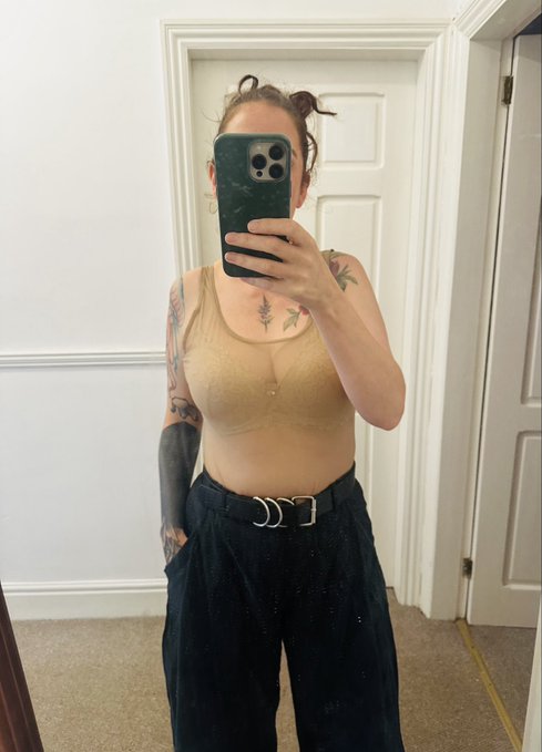 A mirror selfie of a person with tattoos wearing a tan crop top and black high-waisted pants with a statement buckle belt.
