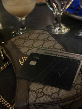 A black American Express (Amex) Platinum card rests on a Gucci monogram bag on a table, with a drink and glassware visible in the background.