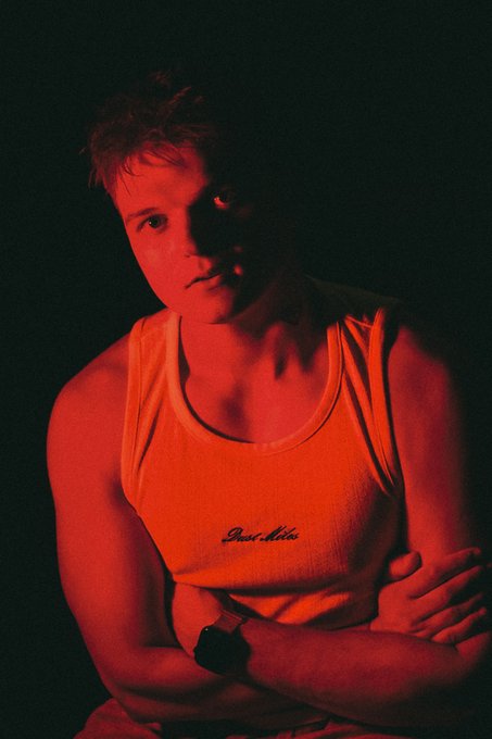 A young man in an orange "Deal Affair" tank top stands with arms crossed under dramatic red lighting, his serious expression and pose conveying a bold, moody aesthetic that complements the stylized "LPHA" branding of the post.