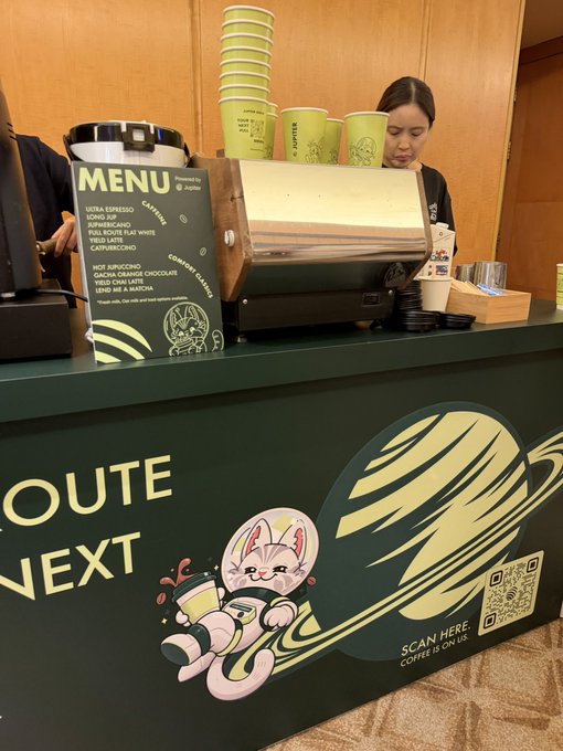 Joffre's coffee shop counter with a green branded front featuring a cute cat mascot, a menu board listing drinks like nitro espresso and hot cappuccino, and an employee behind the counter with stacks of green paper cups.