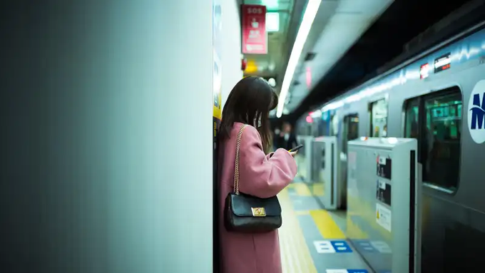 A woman in a pink coat standing on a train platform, looking at her phone with a black handbag over her shoulder.