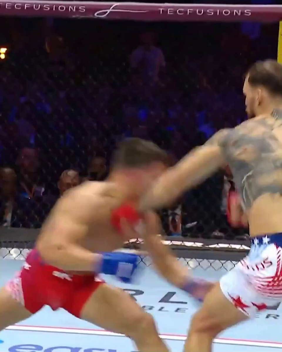 The image shows two men fighting each other, with one man wearing red shorts and a white shirt while the other has blue shorts and a black tank top. The fighter in the red shorts appears to be landing a punch at his opponent who is holding up his fist. The background of the image reveals that there are people watching the fight from behind a fence or cage, indicating it might be taking place in an arena or a similar setting.
